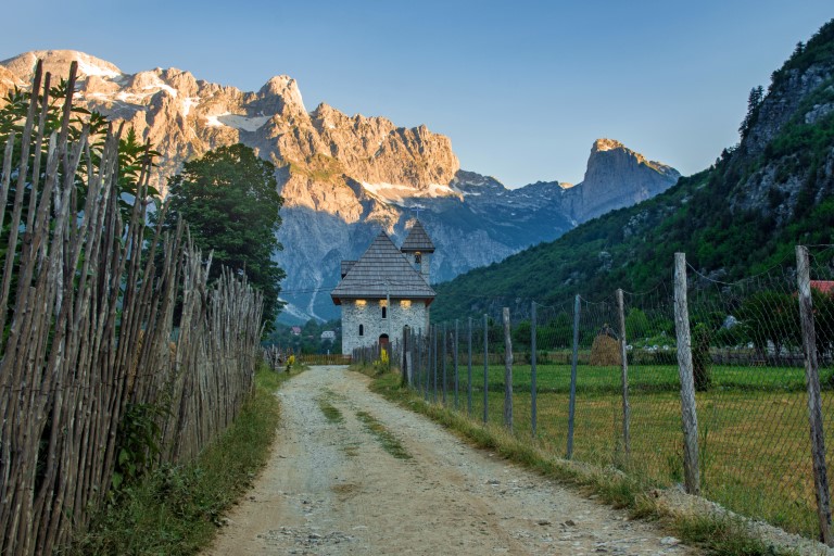 a path with wooden fence leads the catholic church in the Theth Valley, on background the mountains of Albanian Alps